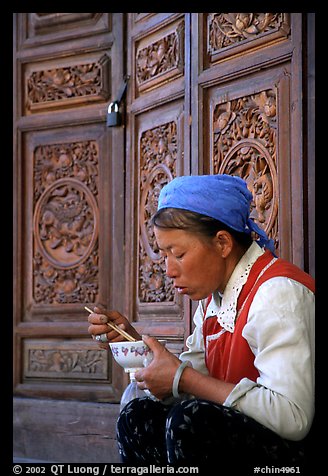 Bai woman eating from a bowl in front of carved wooden doors. Dali, Yunnan, China (color)