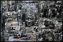 Boy sits next to large stone smiling faces, the Bayon. Angkor, Cambodia (color)