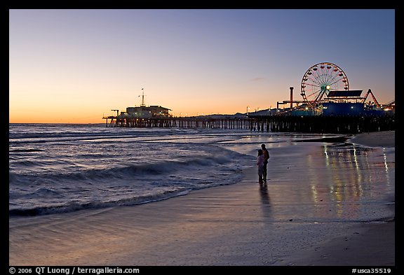 Couple reflected in wet sand at sunset, with pier and Ferris Wheel behind. Santa Monica, Los Angeles, California, USA