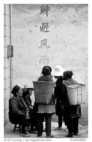 Elderly women with back baskets in front of a wall with Chinese scripture. Shaping, Yunnan, China