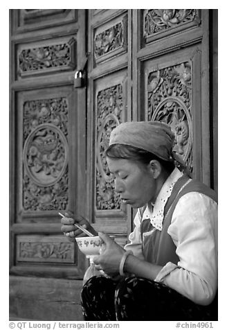 Bai woman eating from a bowl in front of carved wooden doors. Dali, Yunnan, China