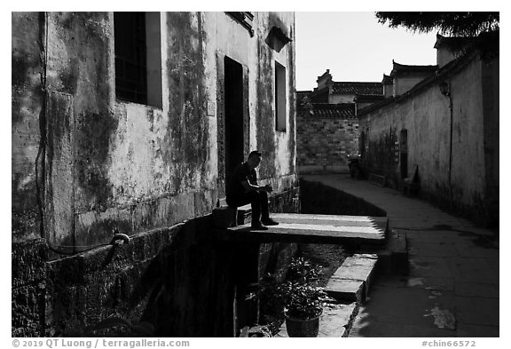 Man sitting in front of house on bridge over stream. Xidi Village, Anhui, China (black and white)