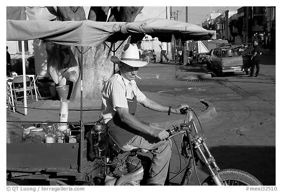 Man with cigarette riding a motorcycle-powered food stand on town plaza. Mexico (black and white)