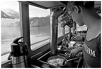 Woman prepares breakfast eggs aboard small tour boat, with glacier in view. Glacier Bay National Park, Alaska, USA. (black and white)