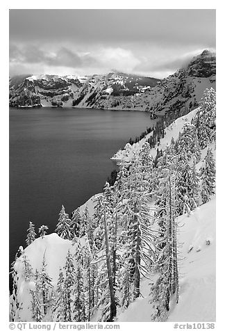 Cliffs, conifer trees, and lake in winter with cloudy skies. Crater Lake National Park, Oregon, USA.