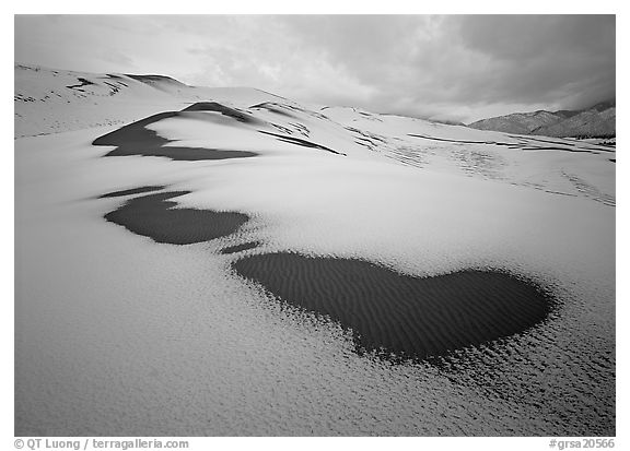 Patches of uncovered sand in snow-covered dunes, mountains, and dark clouds. Great Sand Dunes National Park and Preserve (black and white)
