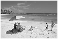 Couple sitting under sun unbrella with children playing around, Poipu Beach, mid-day. Kauai island, Hawaii, USA (black and white)