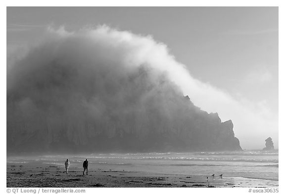 Couple walking on the beach, with Morro Rock and fog behind. Morro Bay, USA (black and white)