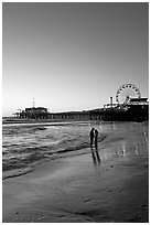 Couple standing on the beach at sunset, with pier and Ferris Wheel behind. Santa Monica, Los Angeles, California, USA (black and white)