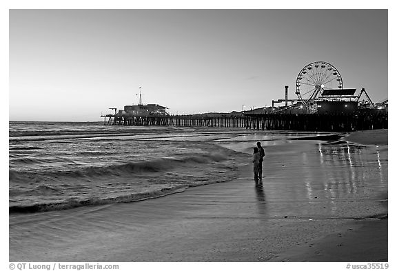 Couple reflected in wet sand at sunset, with pier and Ferris Wheel behind. Santa Monica, Los Angeles, California, USA