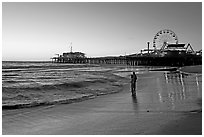 Couple reflected in wet sand at sunset, with pier and Ferris Wheel behind. Santa Monica, Los Angeles, California, USA (black and white)