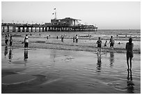 Beachgoers near Santa Monica Pier reflected in wet sand, sunset. Santa Monica, Los Angeles, California, USA (black and white)