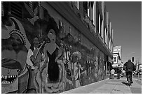 Man riding bicycle on sidewalk past mural, Mission District. San Francisco, California, USA ( black and white)