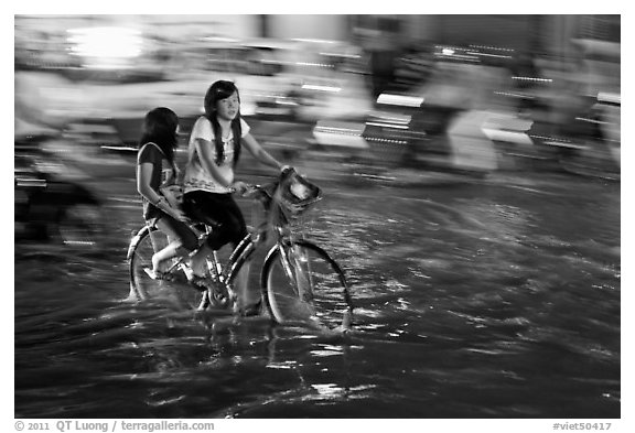 Girls sharing night bicycle ride through water of flooded street. Ho Chi Minh City, Vietnam (black and white)