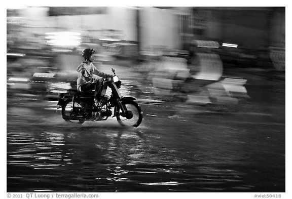 Motorcyclist speeding on wet street at night, with streaks giving sense of motion. Ho Chi Minh City, Vietnam (black and white)