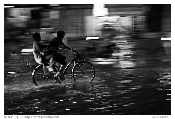 Men sharing bicycle ride at night on wet street. Ho Chi Minh City, Vietnam (black and white)