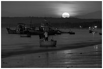 Fisherman paddling on coracle boat towards fishing boats at moonset. Mui Ne, Vietnam (black and white)