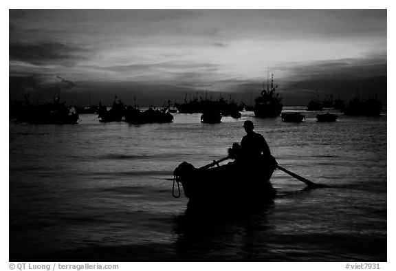 Man in a small boat, with moored boats seen against a vivid sunset. Vung Tau, Vietnam (black and white)