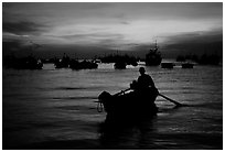 Man in a small boat, with moored boats seen against a vivid sunset. Vung Tau, Vietnam (black and white)