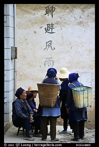 Elderly women with back baskets in front of a wall with Chinese scripture. Shaping, Yunnan, China