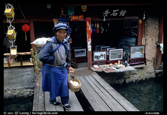 Naxi woman peddling eggs  to local residents walks acros a canal. Lijiang, Yunnan, China