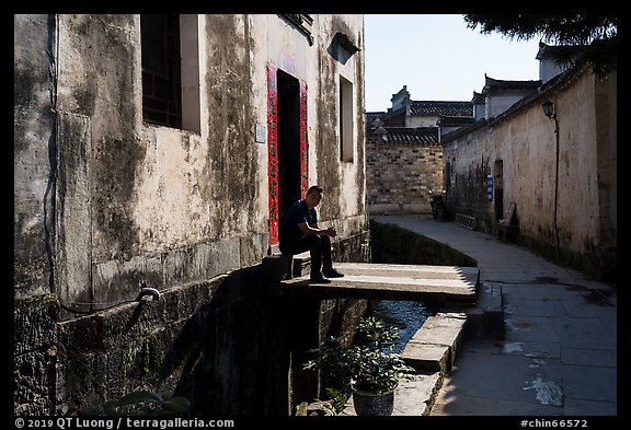 Man sitting in front of house on bridge over stream. Xidi Village, Anhui, China (color)