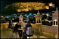 Travelers on rooftop terrace with view of Mehrangarh Fort by night. Jodhpur, Rajasthan, India (color)