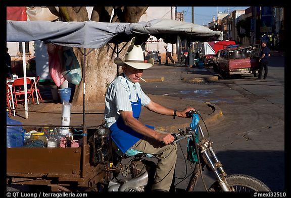 Man with cigarette riding a motorcycle-powered food stand on town plaza. Mexico (color)