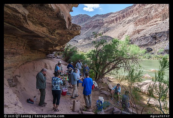 Group of rafters during a meal on a sandy ledge beside the Colorado River, shaded by rock overhangs. Grand Canyon National Park, Arizona (color)