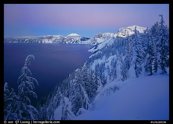 Snow-covered rim and trees, lake and mountains, dusk. Crater Lake National Park, Oregon, USA.
