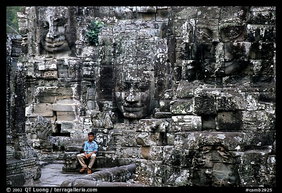 Boy sits next to large stone smiling faces, the Bayon. Angkor, Cambodia (color)