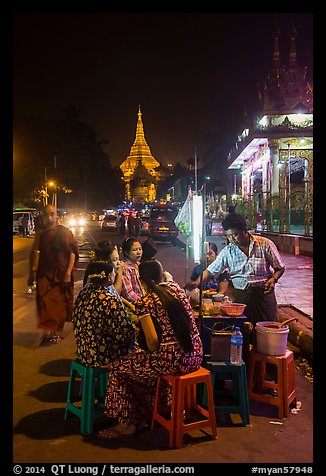 Women eating at street stall at night with Shwedagon Pagoda in background. Yangon, Myanmar (color)