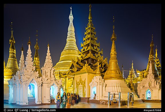 Women walking, stupas, shrines, and Main Stupa at night, Shwedagon Pagoda. Yangon, Myanmar