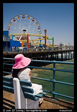 Woman sitting on bench with pink hat and ferris wheel. Santa Monica, Los Angeles, California, USA (color)