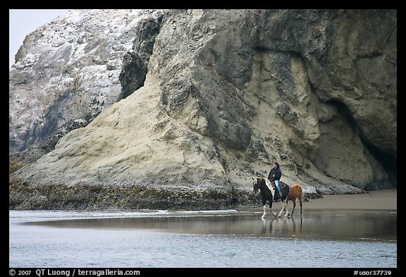 Woman horse-riding on beach next to sea cave entrance. Bandon, Oregon, USA