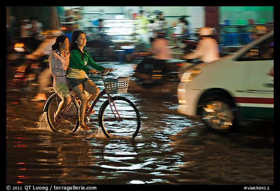 Women sharing a bicycle ride at night on a water-filled street. Ho Chi Minh City, Vietnam (color)
